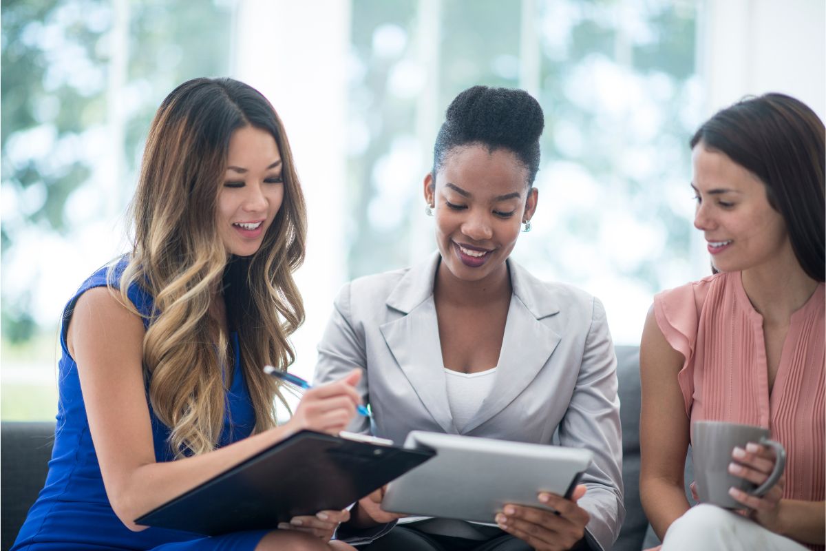 Three women in professional attire are sitting together. One is holding a clipboard, another a tablet, and the third a cup as they engage in a discussion about their next nurture campaign.