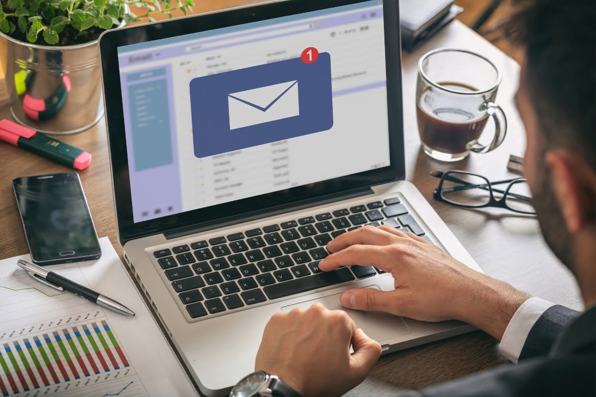 A person using a laptop receives an email notification pop-up on the screen, indicating one new email. A coffee cup, smartphone, and documents are on the desk, highlighting a moment where email targeting meets daily productivity.