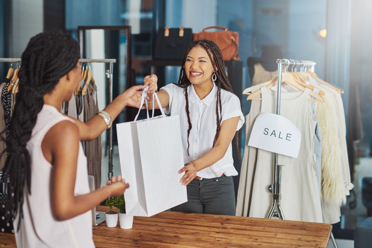 In a clothing store with a sale sign in the background, one woman hands a shopping bag to another, potentially illustrating how to use geofencing marketing strategies effectively.