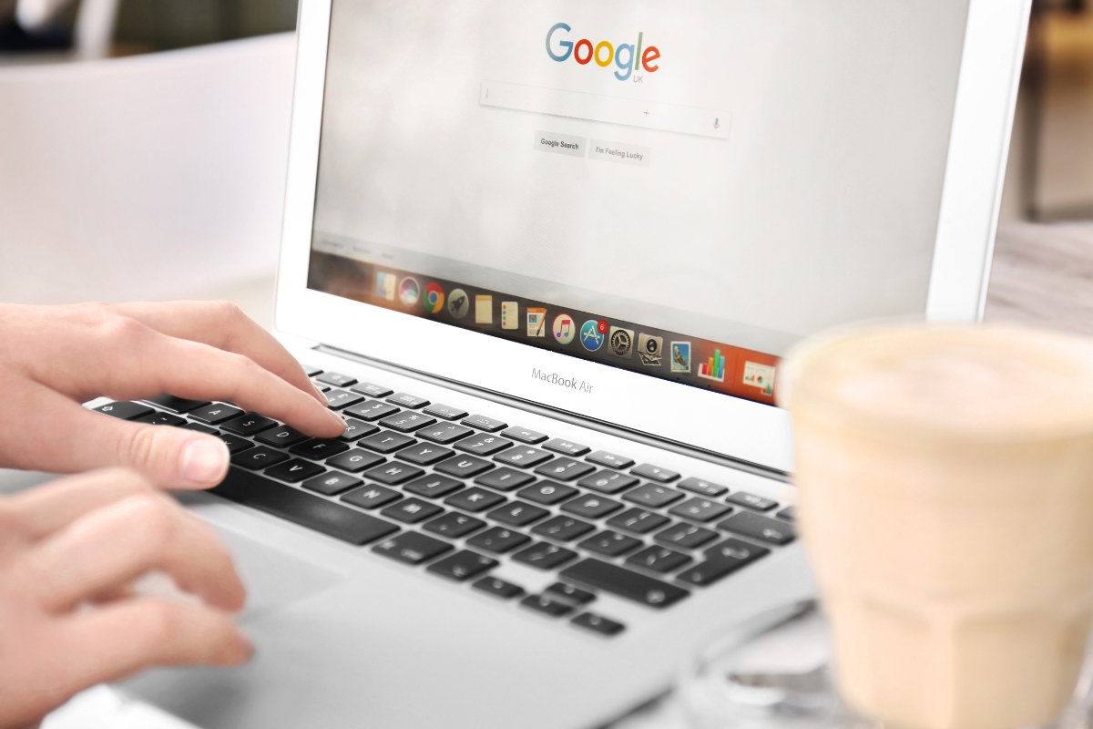 A person types on a MacBook Air with a Google search page open, looking up "what is Google Workspace." A coffee cup sits on the table next to the laptop.