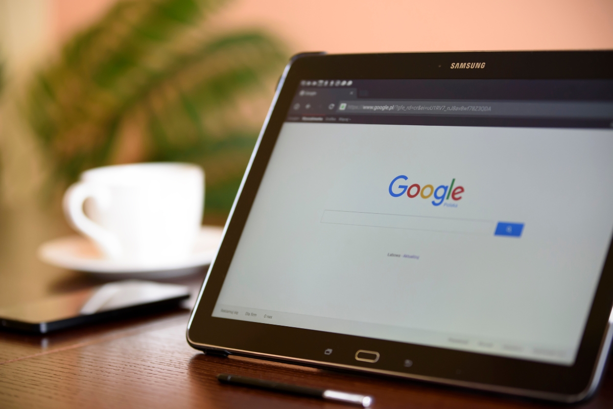 A Samsung tablet displaying the Google search homepage is placed on a table, subtly hinting at queries like "what is Google Workspace?" A cup and a blurred plant are in the background.