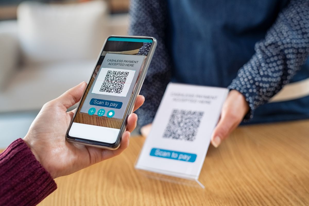 A person's hand holding a smartphone scanning a QR code for payment from a table while another person holds a stand displaying the code, making one wonder, what are QR codes?