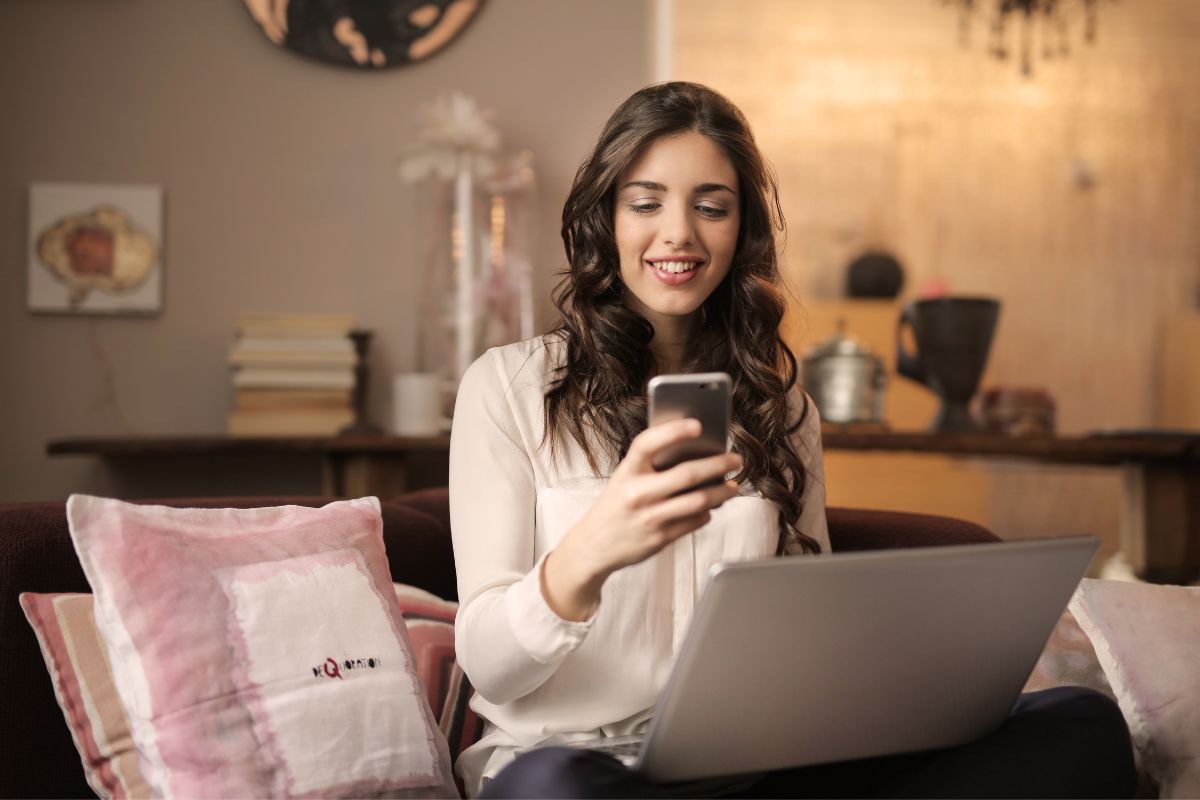 A woman sitting on a couch uses her smartphone while a laptop rests on her lap, possibly researching what are SERPs. She is in a comfortable living room with a tidy background.