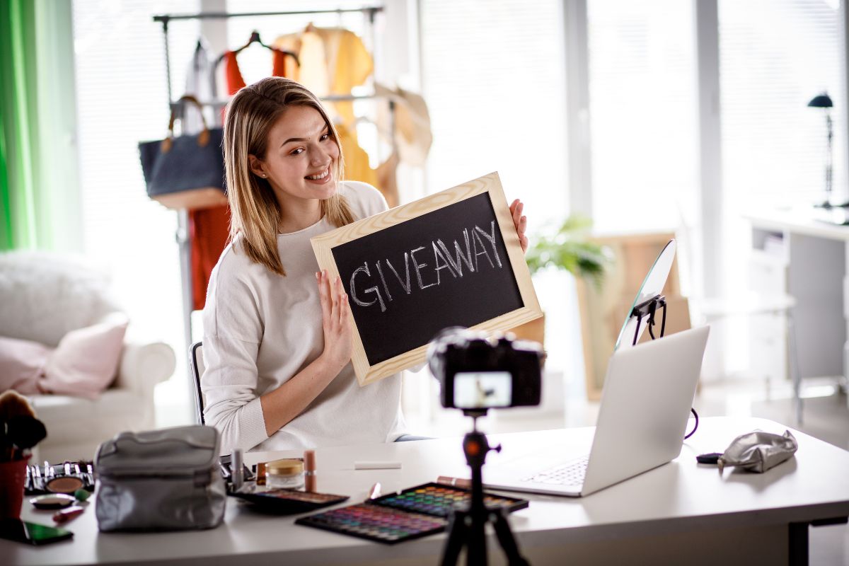 A woman with light brown hair holds a chalkboard sign reading "Giveaway," while sitting at a desk with a laptop, makeup items, and a camera recording her. She is explaining what is a lead magnet and how it can boost your online presence.