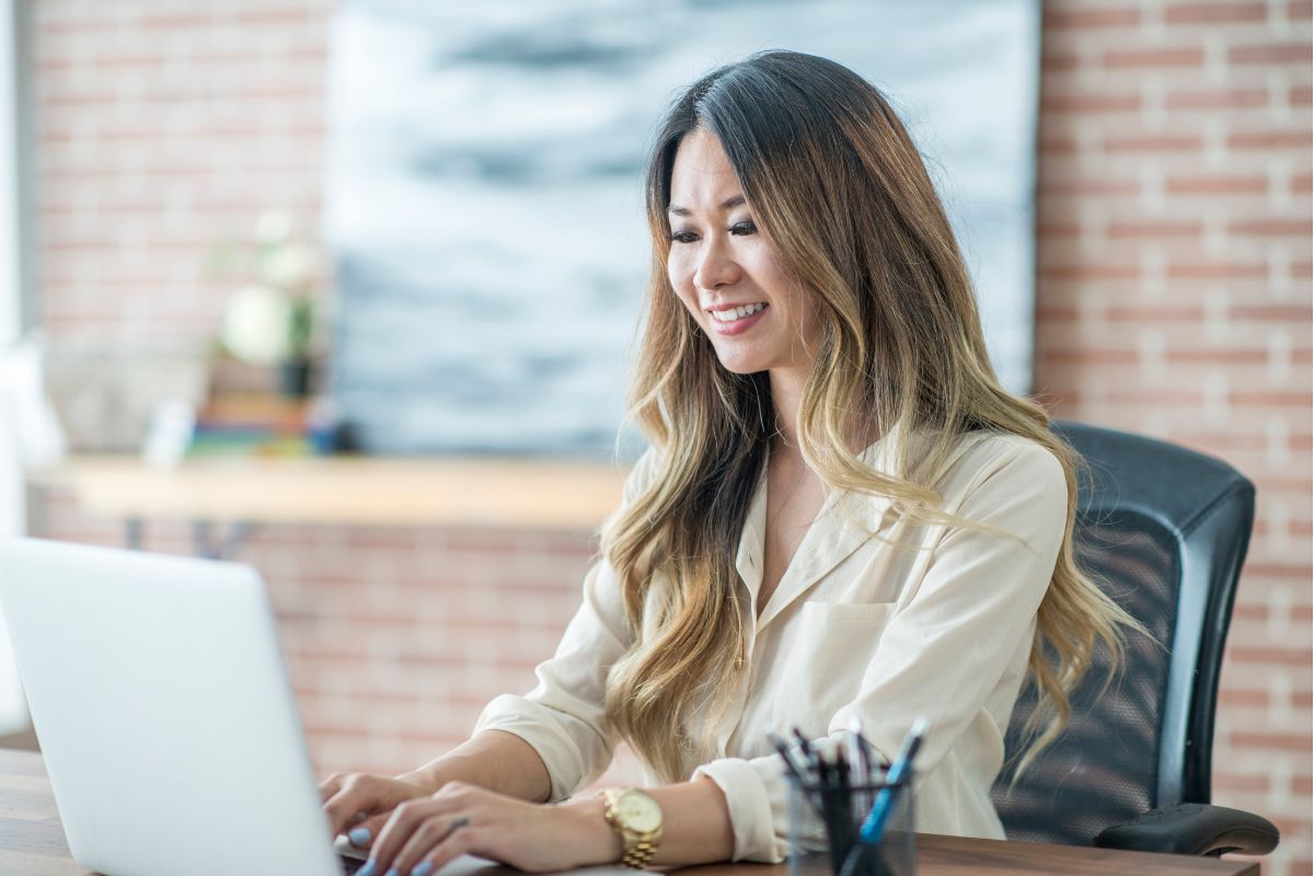 A woman with long hair works on a laptop at a desk in an office setting, possibly researching how to create an email signature.