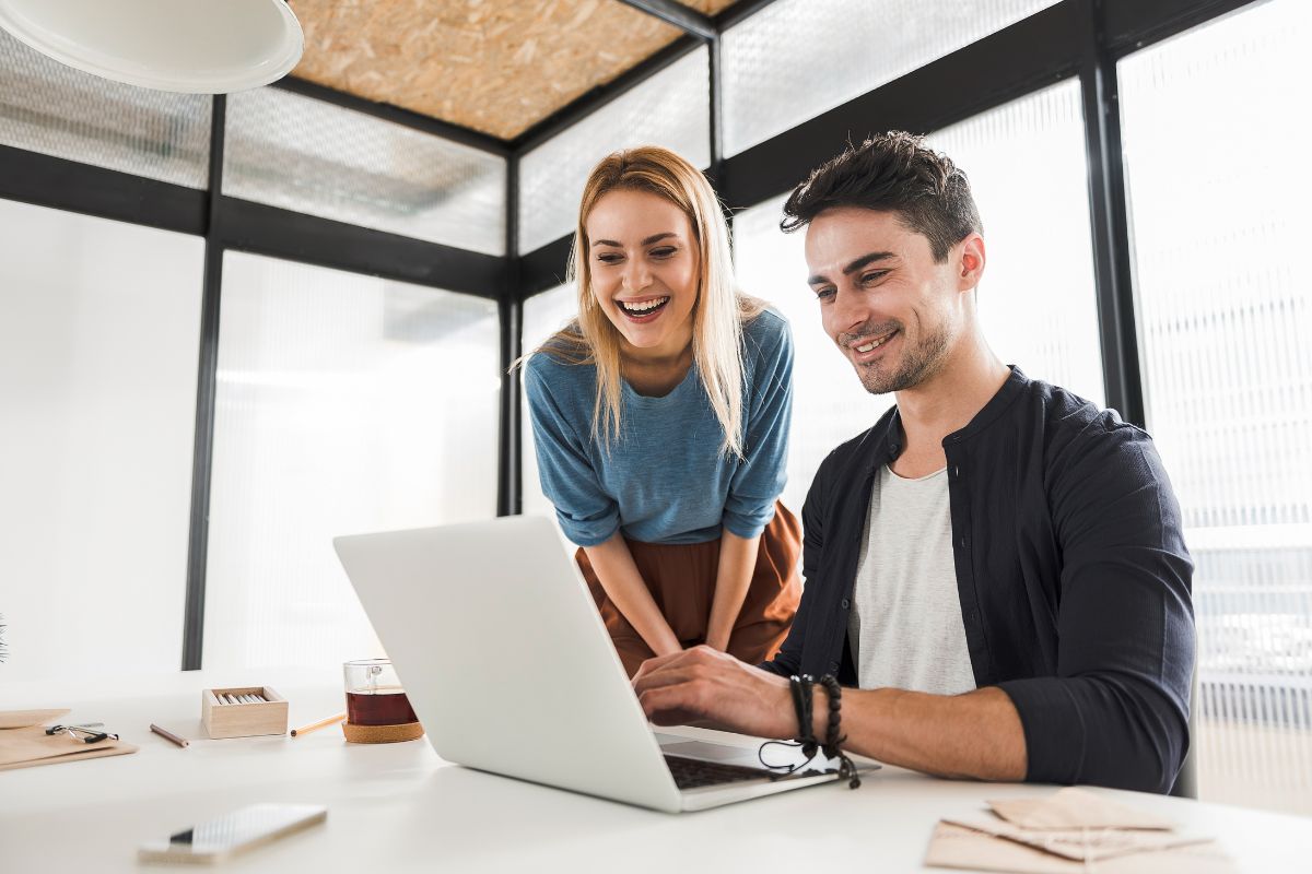 Two people are smiling while looking at a laptop in an office setting. The person sitting is typing, creating a new landing page, and the person standing is leaning in to observe, aiming to boost conversions.