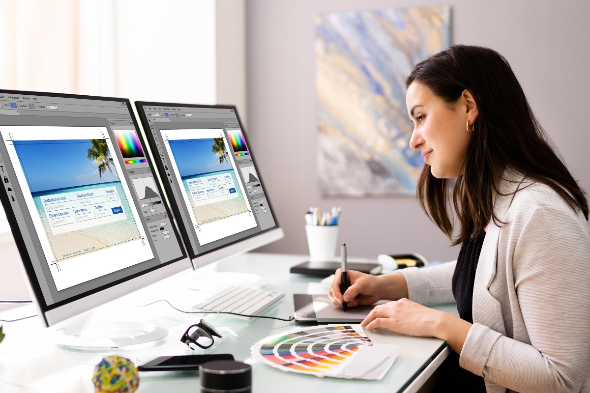 A woman sits at a desk working on graphic design using two monitors and a digital pen tablet, with color swatches and office supplies around her, creating visuals for how to make a website for a business.