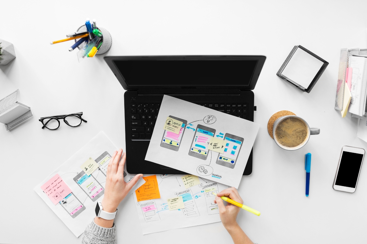 An overhead view of a person working on UX design drafts placed on a laptop keyboard, potentially pondering how to make a website for a business. Nearby are a coffee cup, smartphone, glasses, and various office supplies.