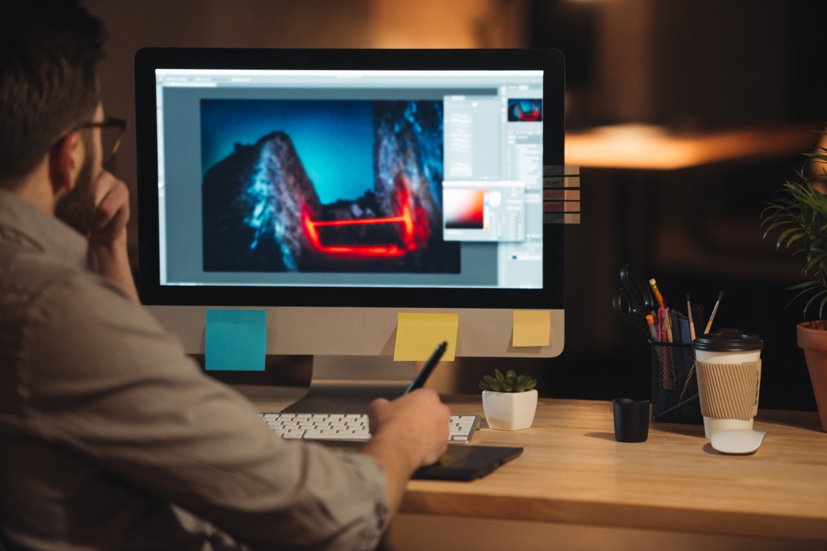 A person working on photo editing software on a large monitor, with various tools and options displayed. The desk contains sticky notes about how to make a website for a business, a potted plant, pens, and a coffee cup.