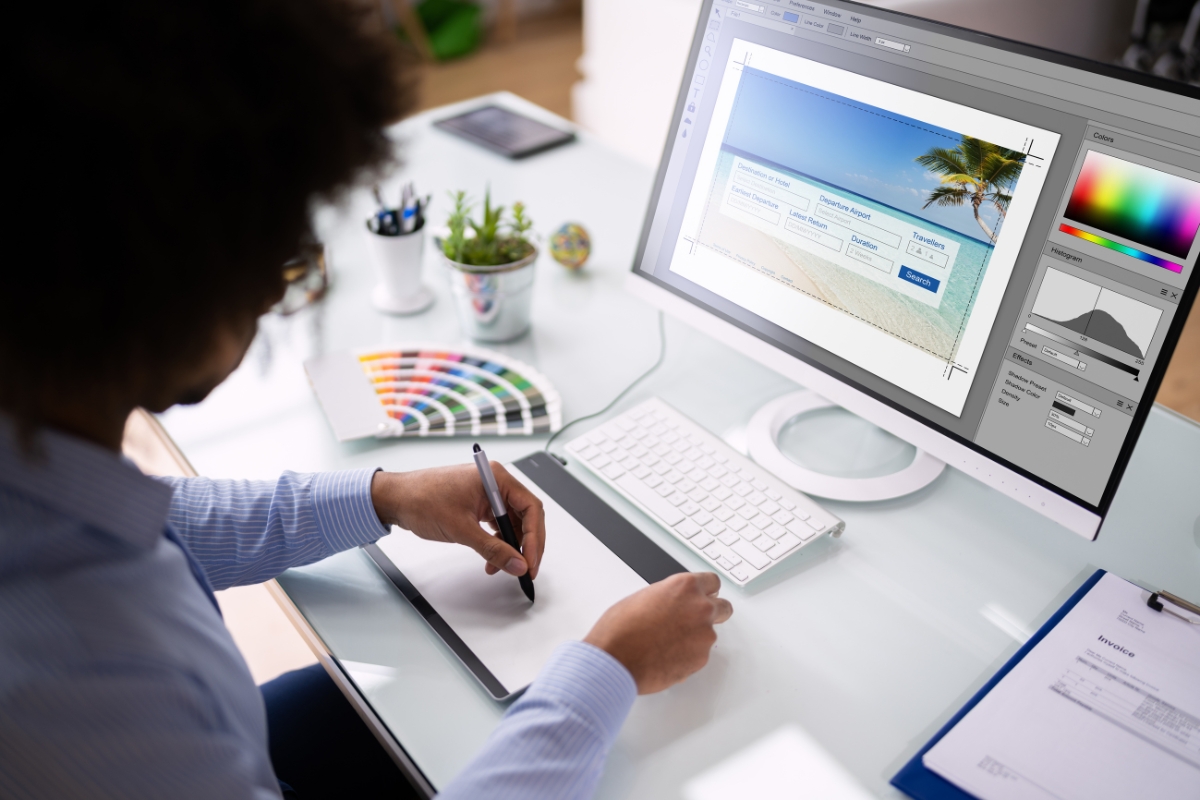 Person working on graphic design at a desk with a digital pen tablet, large computer monitor, colorful palette, and various desk accessories, while considering how to make a website for a business.