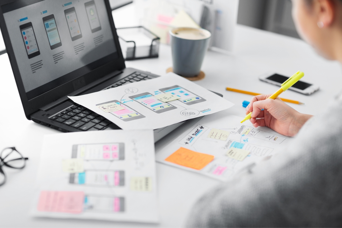 A person is working on UX design at a desk with a laptop, focusing on how to make a website for a business. They are using a highlighter on printed wireframes while additional wireframes are displayed on the laptop screen. A cup and stationery are nearby.