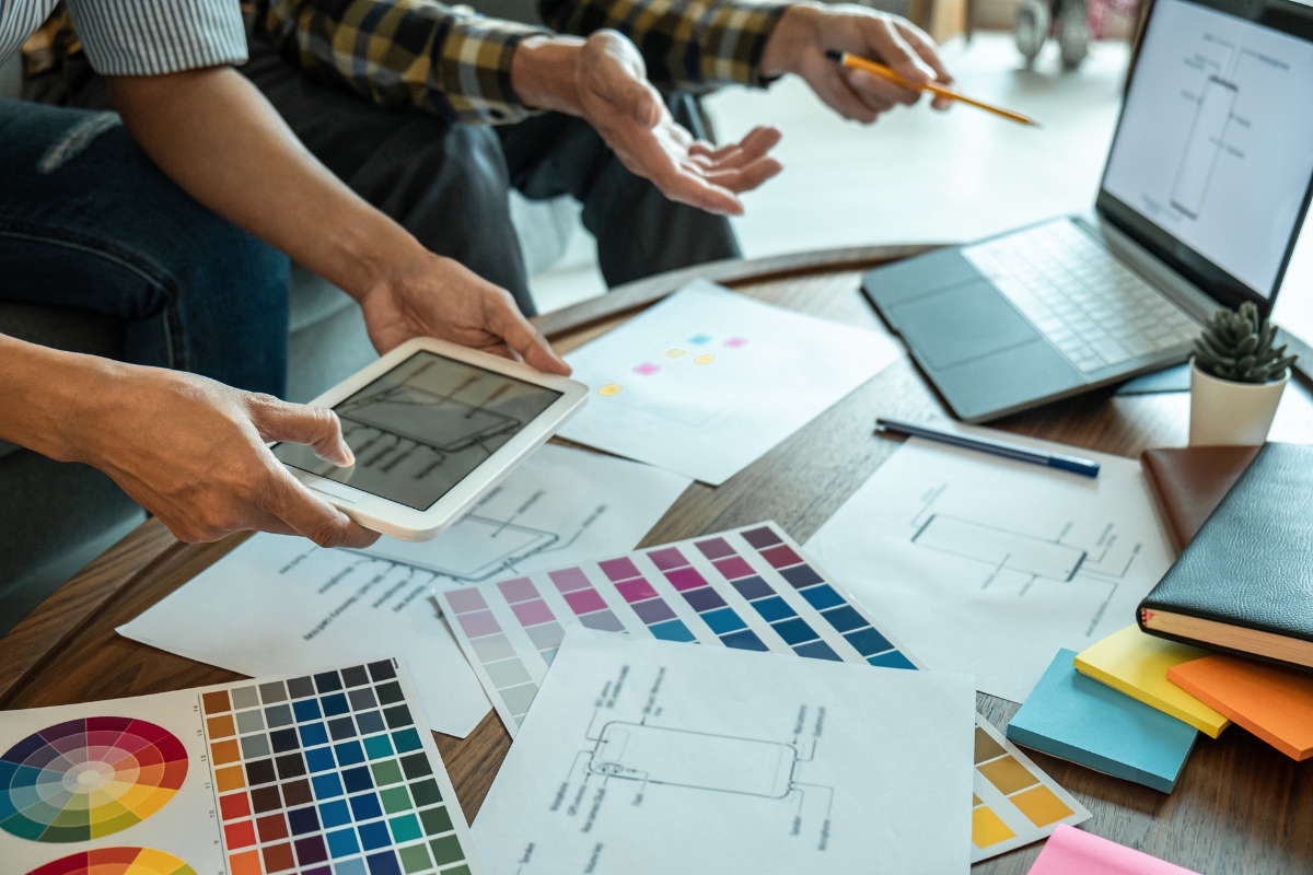 Two people discussing how to make a website for a business over a table filled with color swatches, printed diagrams, a tablet, a laptop, notepads, and sticky notes. One is pointing with a pencil while the other holds the tablet.