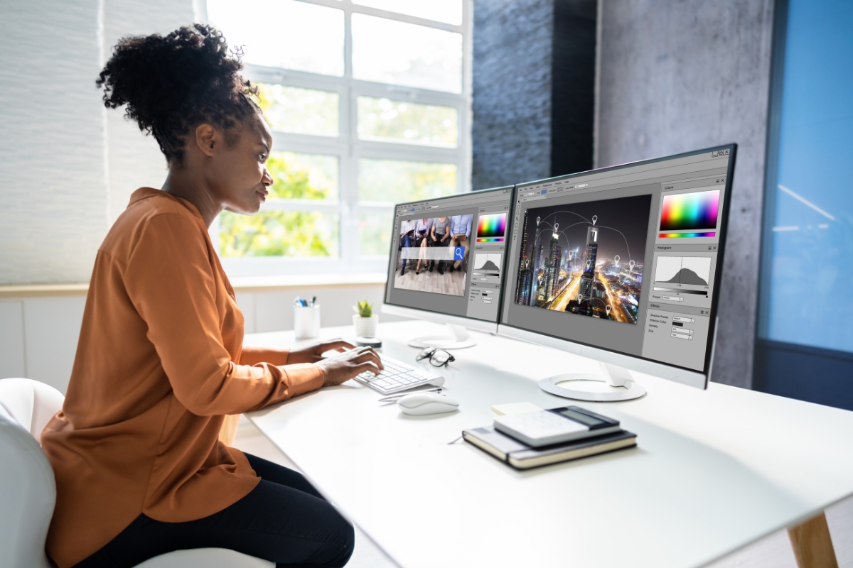 A person sits at a desk working on a dual-monitor setup, editing images using graphic design software while researching how to make a website for a business. The workspace includes a keyboard, mouse, notebooks, and a cup on a bright, clean desk.