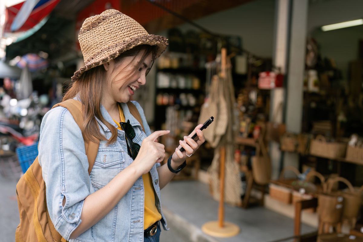 A woman wearing a hat and backpack smiles while using her phone. She stands in front of a shop, perhaps reading about how to use geofencing marketing.