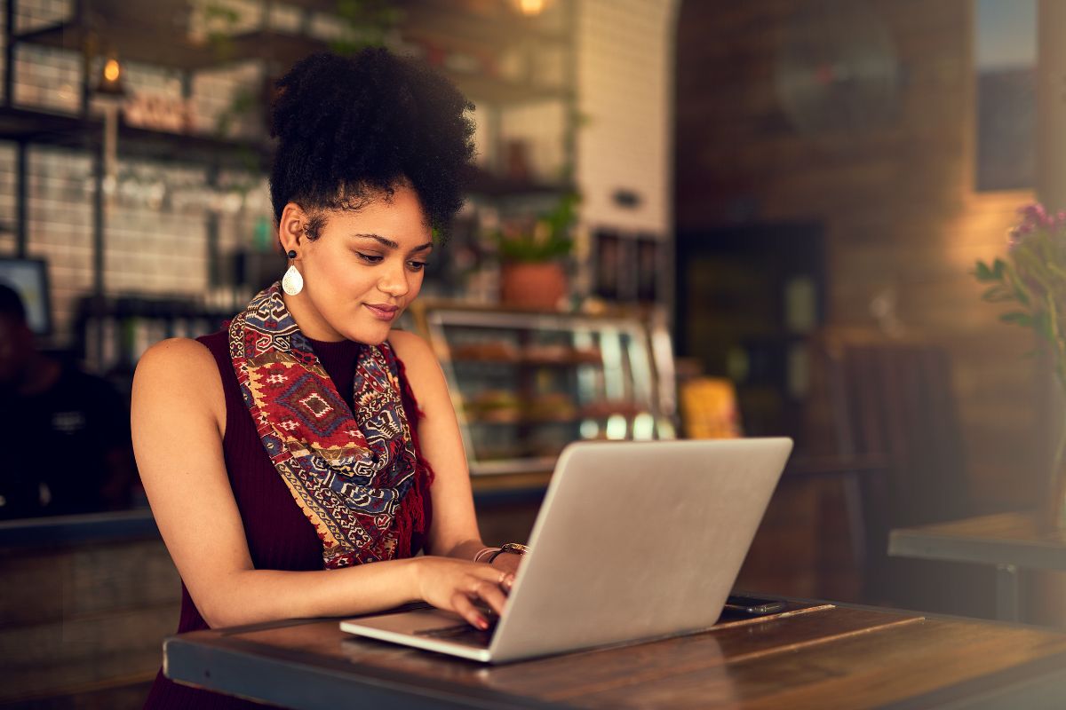 A woman types on a laptop while seated at a wooden table in a cozy café, possibly drafting "How To Write A Blog Post." She wears a patterned scarf and earrings, with shelves and a counter visible in the background.