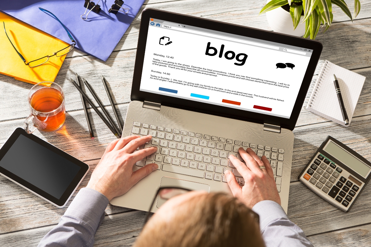 Person typing on a laptop displaying a blog webpage, surrounded by stationery, a calculator, a cup of tea, and a tablet on a wooden desk. The scene captures the essence of learning how to write quality content in an organized, productive workspace.