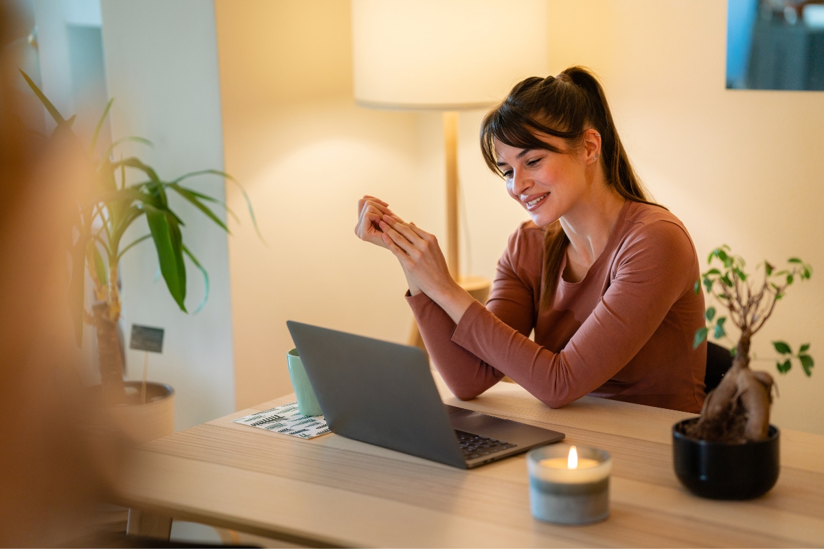 A woman sits at her desk, holding a phone and smiling, with a laptop open beside her. A potted plant adds a touch of nature, while a lit candle creates a cozy ambiance. She appears to be researching how to write quality content.