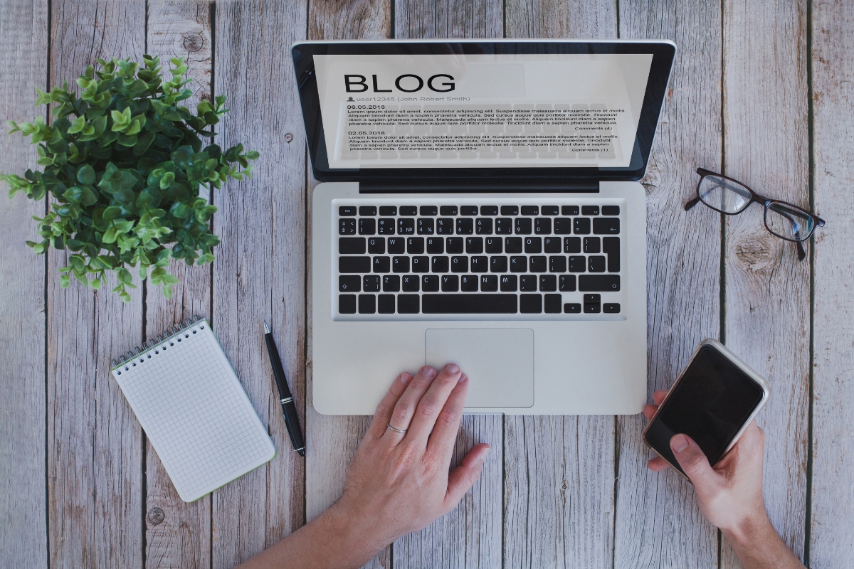 A person is using a laptop with a "BLOG" page open, possibly researching how to write quality content. A plant, notepad, pen, glasses, and smartphone are also on the wooden table.