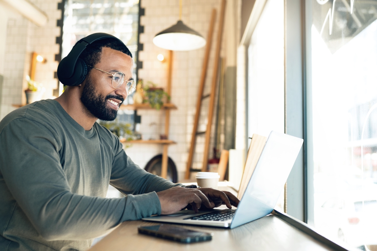 A person wearing headphones and glasses works on a laptop at a cafe, perhaps researching how to write quality content, with a smartphone placed on the table in front of them.