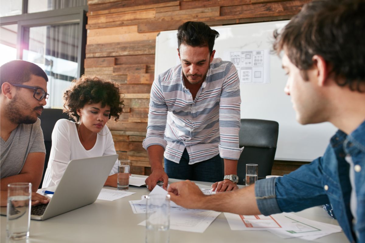 Four people are gathered around a table in a meeting room. One person is standing and pointing at documents on the table, illustrating the differences between inbound vs outbound marketing, while the others are seated, attentively examining the same documents.