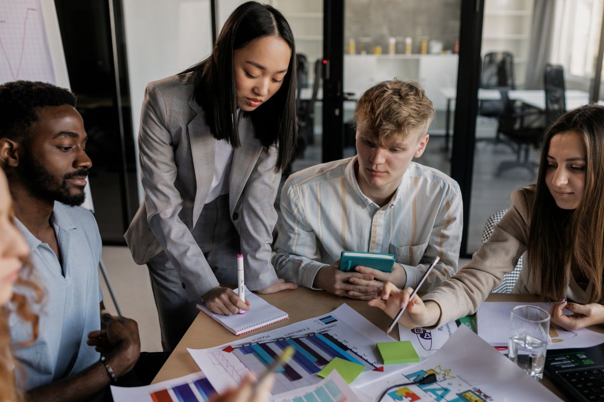 A group of five people collaborating at a table with charts, graphs, and documents. One person is standing, the others are seated, all engaged in a discussion about inbound vs outbound marketing strategies.