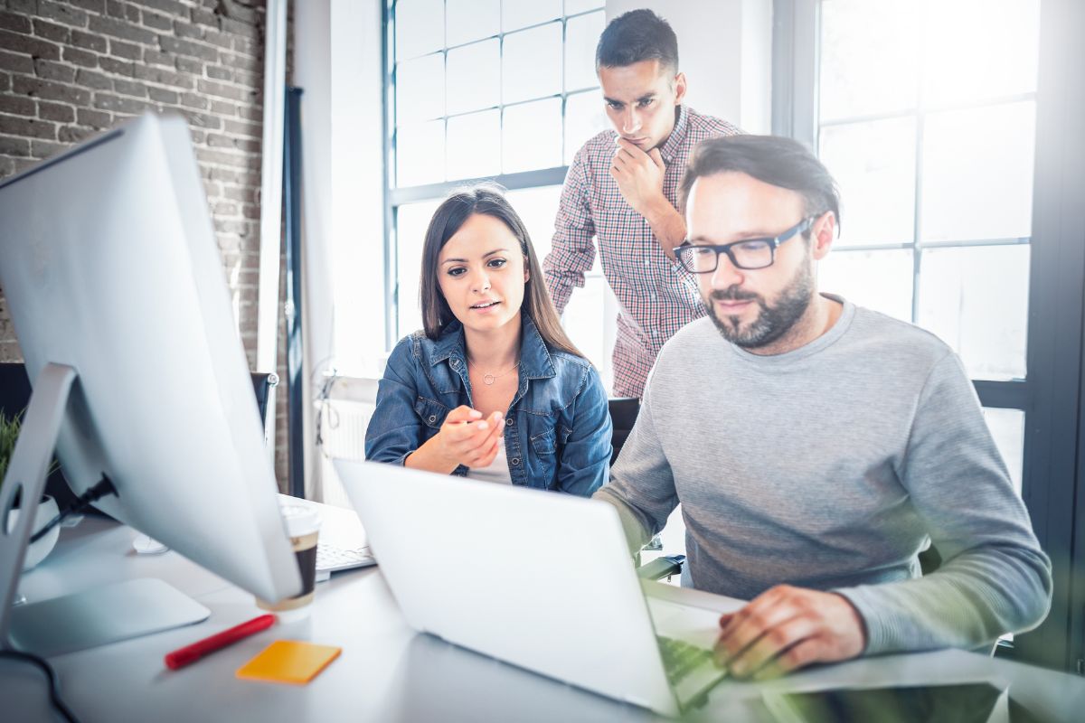 Three people working collaboratively at a desk with two computer screens in a bright office environment, focusing on how to create a landing page that drives conversions quickly.