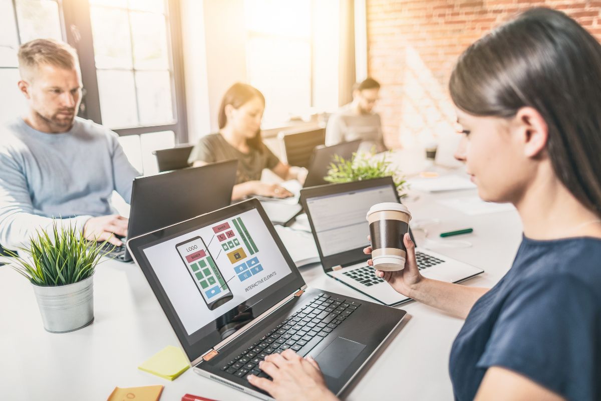 A group of people working on laptops at a shared desk in a bright office. The woman in the foreground holds a coffee cup and focuses on a screen displaying a colorful dashboard titled "How to Create a Landing Page That Drives Conversions Quickly.