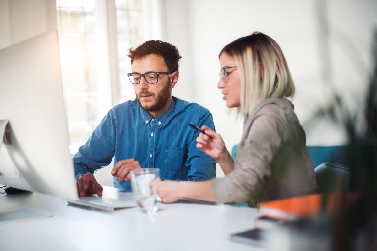 Two people are working together at a desk, looking at a computer screen. The person on the left wears a blue shirt and glasses, while the person on the right has light-colored hair and holds a pen, possibly discussing Inbound vs Outbound Marketing strategies.