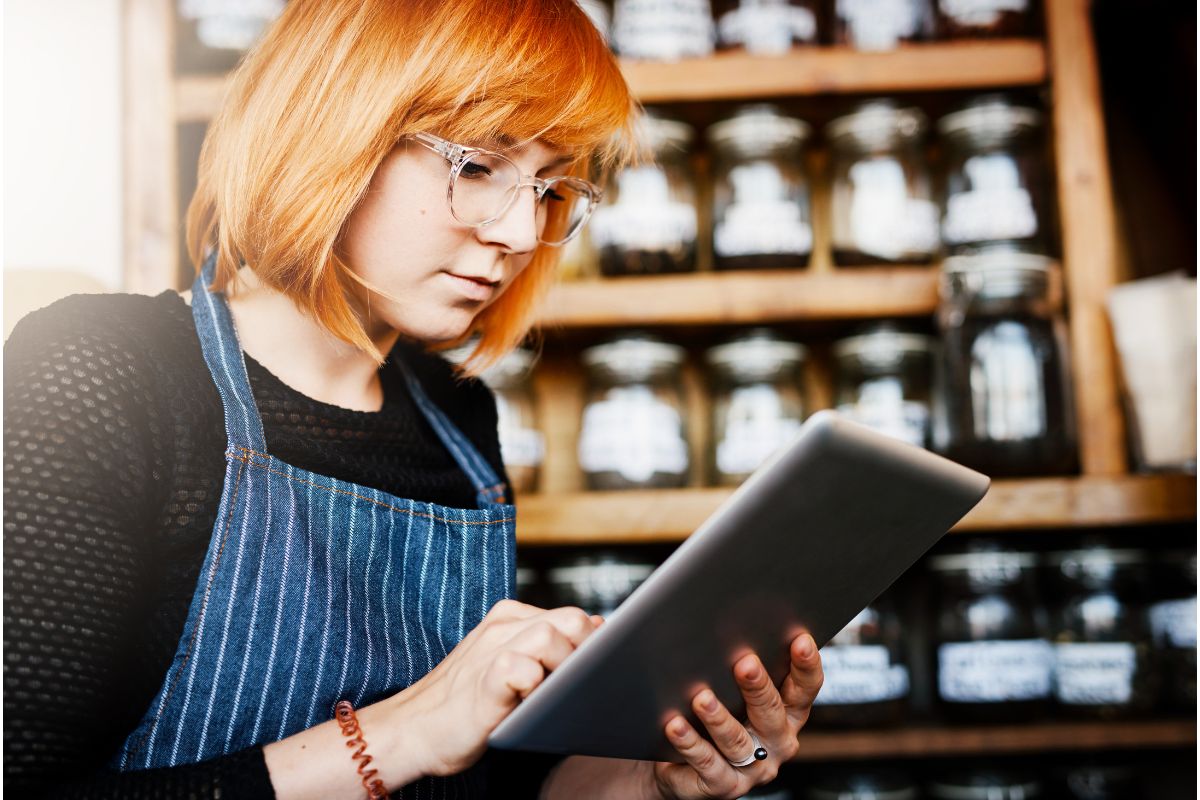 A person with red hair and glasses, wearing an apron, uses a tablet in front of a shelf filled with jars, perhaps researching what is a lead magnet.