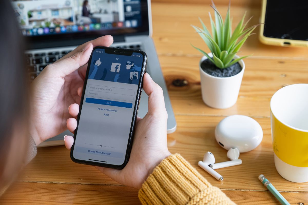 Person holding a smartphone displaying a Facebook login screen over a wooden desk with a laptop, potted plant, earphones, and a cup—an ideal setup for learning how to use social media for business.