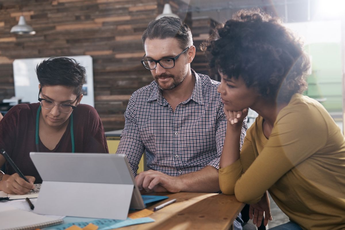 Three individuals are seated at a table, collaborating on a project using a tablet and taking notes, while discussing what is semantic search.