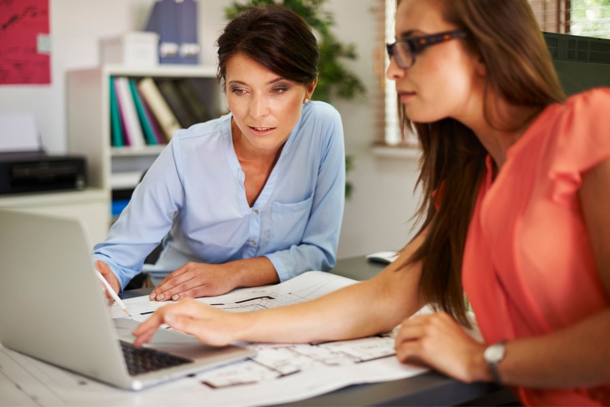 Two women are working together at a desk, one pointing at a laptop screen displaying "what is SEO" while the other reviews printed documents. They appear focused and engaged in their task. Office supplies and shelves are in the background.