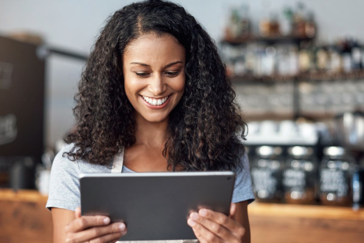 A woman with curly hair and a light-colored shirt smiles while looking at a tablet in a café-like setting, perhaps researching What Are SERPs.