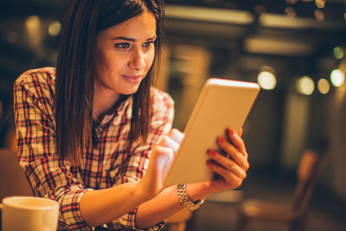 A woman with straight hair in a plaid shirt is sitting at a table and looking at a tablet, possibly reading about "What Are SERPs," with a cup in the foreground and a dimly lit background.