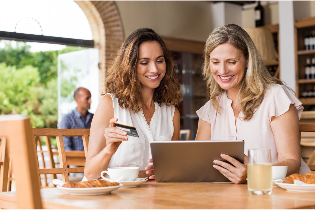 Two women sitting at a table in a café, smiling while looking at a tablet. One woman holds a credit card, possibly about to make an online purchase. There is a cup of coffee, a glass of juice, and pastries on the table beside them. They seem to be enjoying their time together while possibly discussing how to do keyword research.