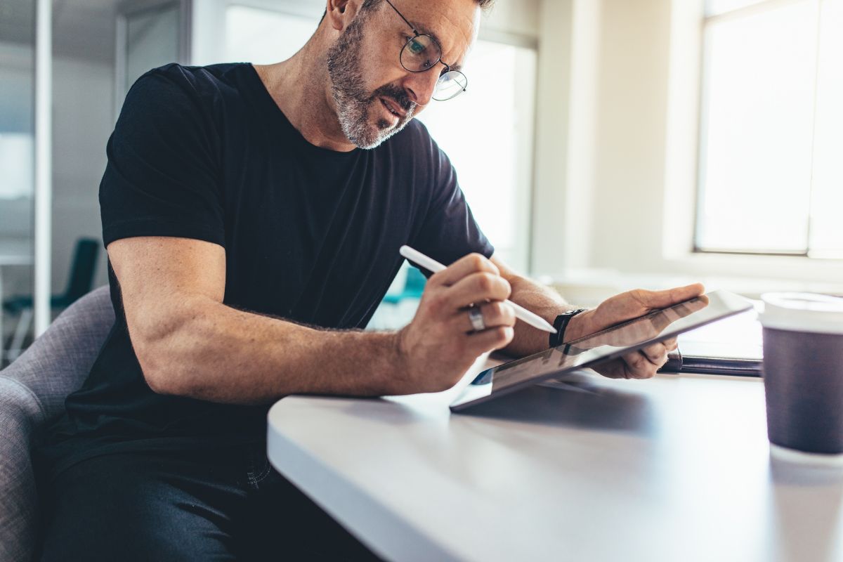 Man with glasses and beard writing on a tablet using a stylus, seated at a table with a cup nearby in a bright room. He appears to be drafting tips on how to create an email signature.