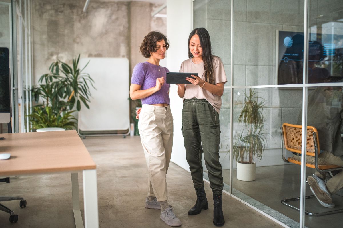 Two individuals stand in an office setting, looking at a tablet together. One person is wearing a purple top and beige pants, and the other is wearing a tan top and green cargo pants. They seem engaged in discussing how to use geofencing marketing strategies for their business.