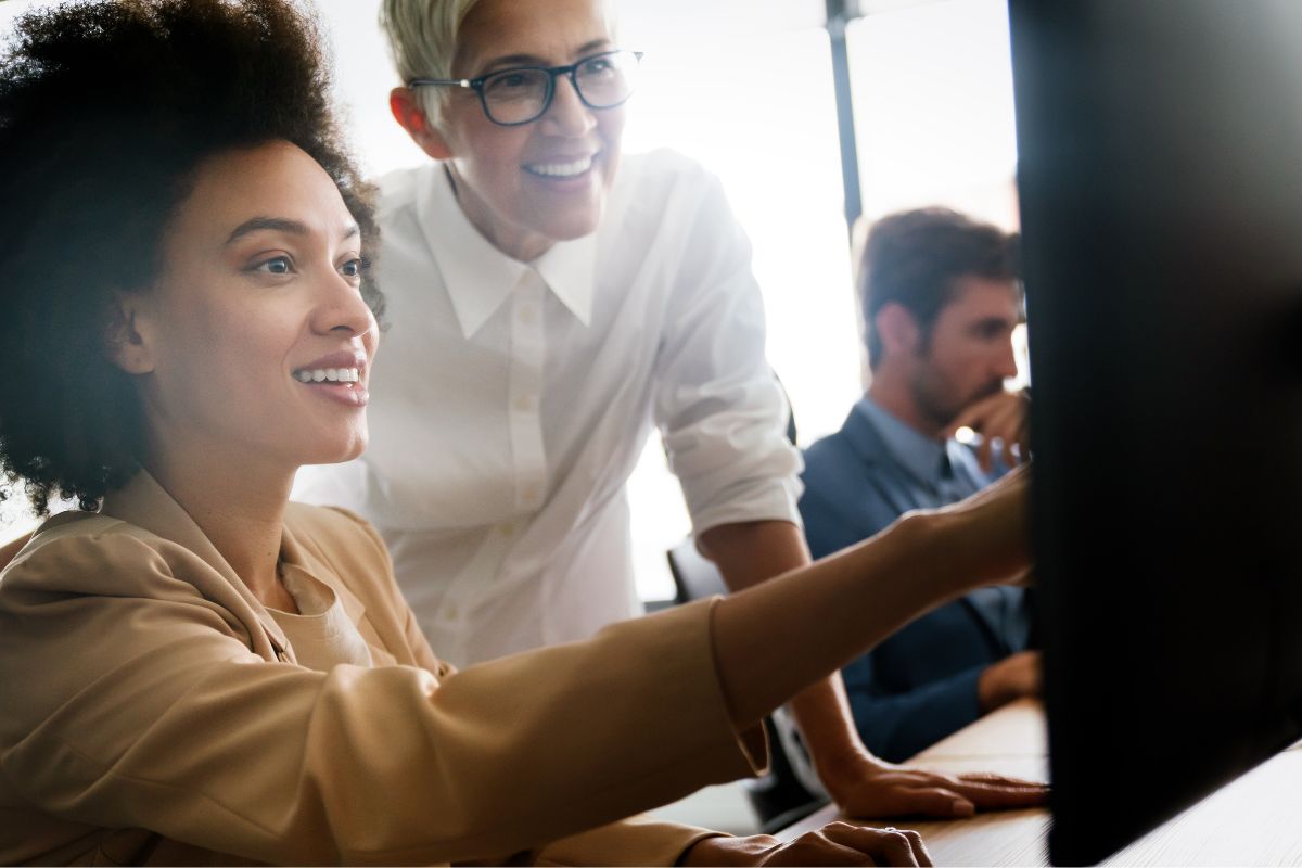 Two women are looking at a computer screen together and smiling, possibly discussing what is NLP. In the background, a man is focused on his computer.