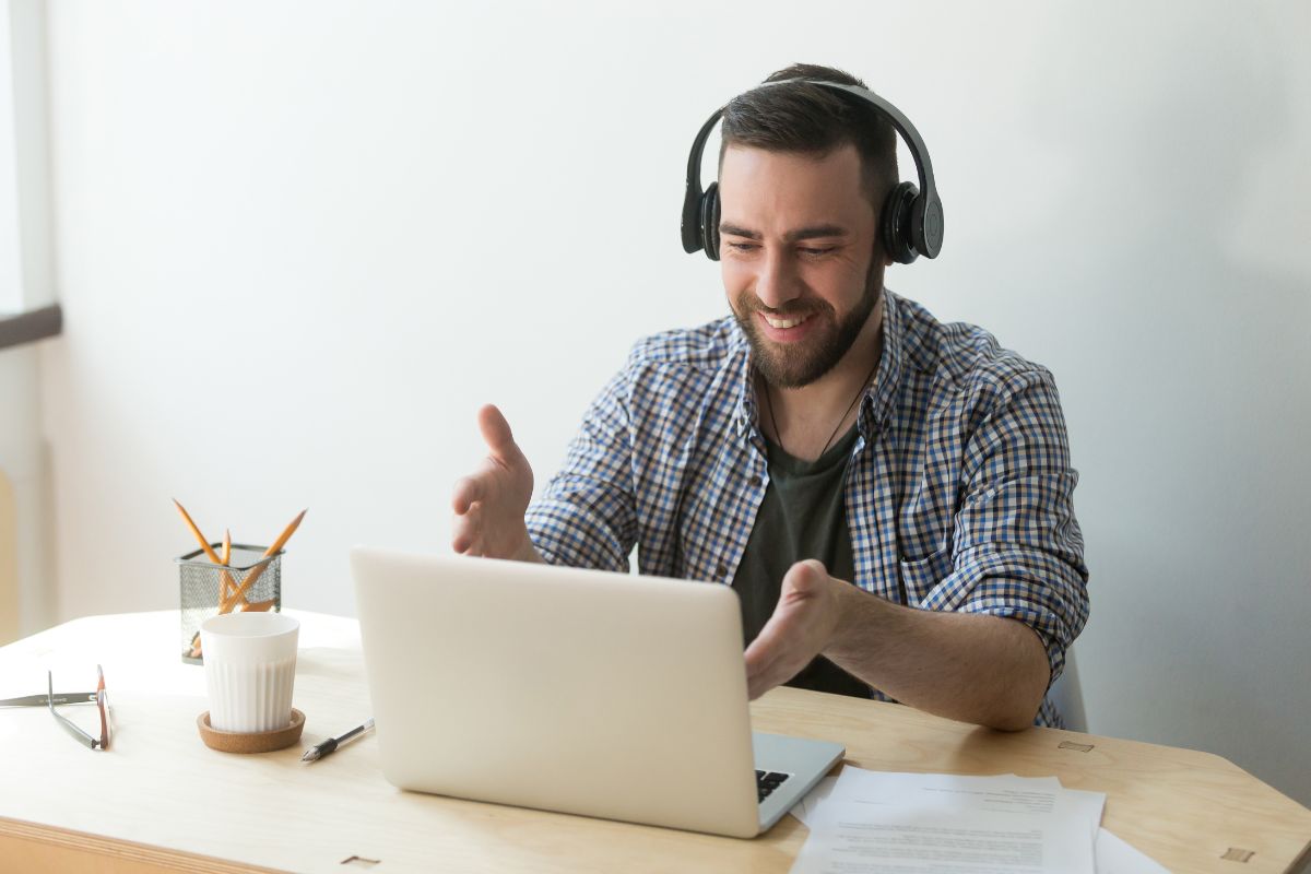 A man wearing headphones sits at a desk, gesturing while looking at his laptop during an online meeting, possibly discussing video testimonials.