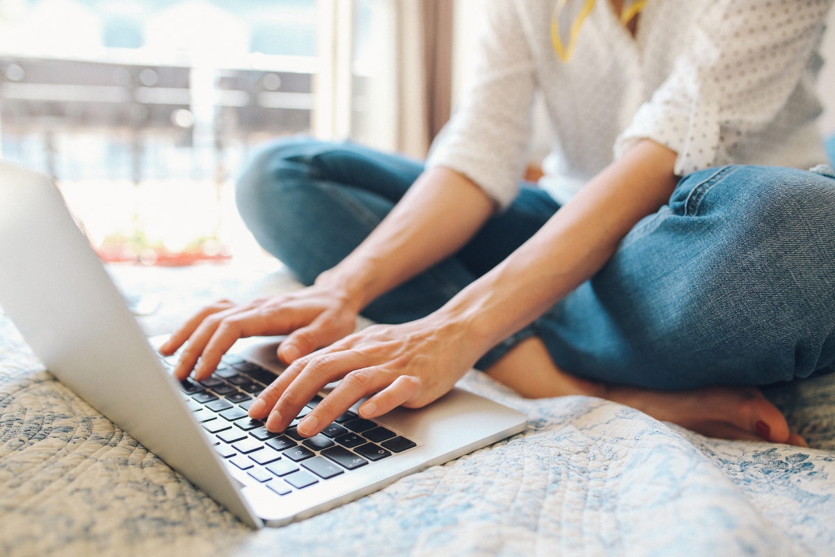 Person sitting cross-legged on a bed, typing on a laptop and likely searching for information like "what is UX writing?" The individual is wearing jeans and a white long-sleeved shirt.