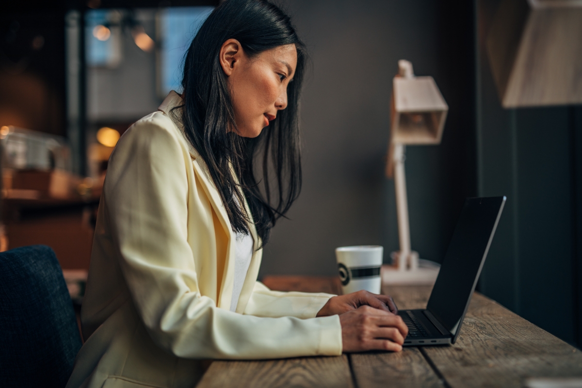 A woman in a yellow blazer works on a laptop at a wooden table in a cafe, with a takeaway coffee cup and a lamp in the background. Curious passersby wonder, "What is UX writing?" as she delves into her screen.