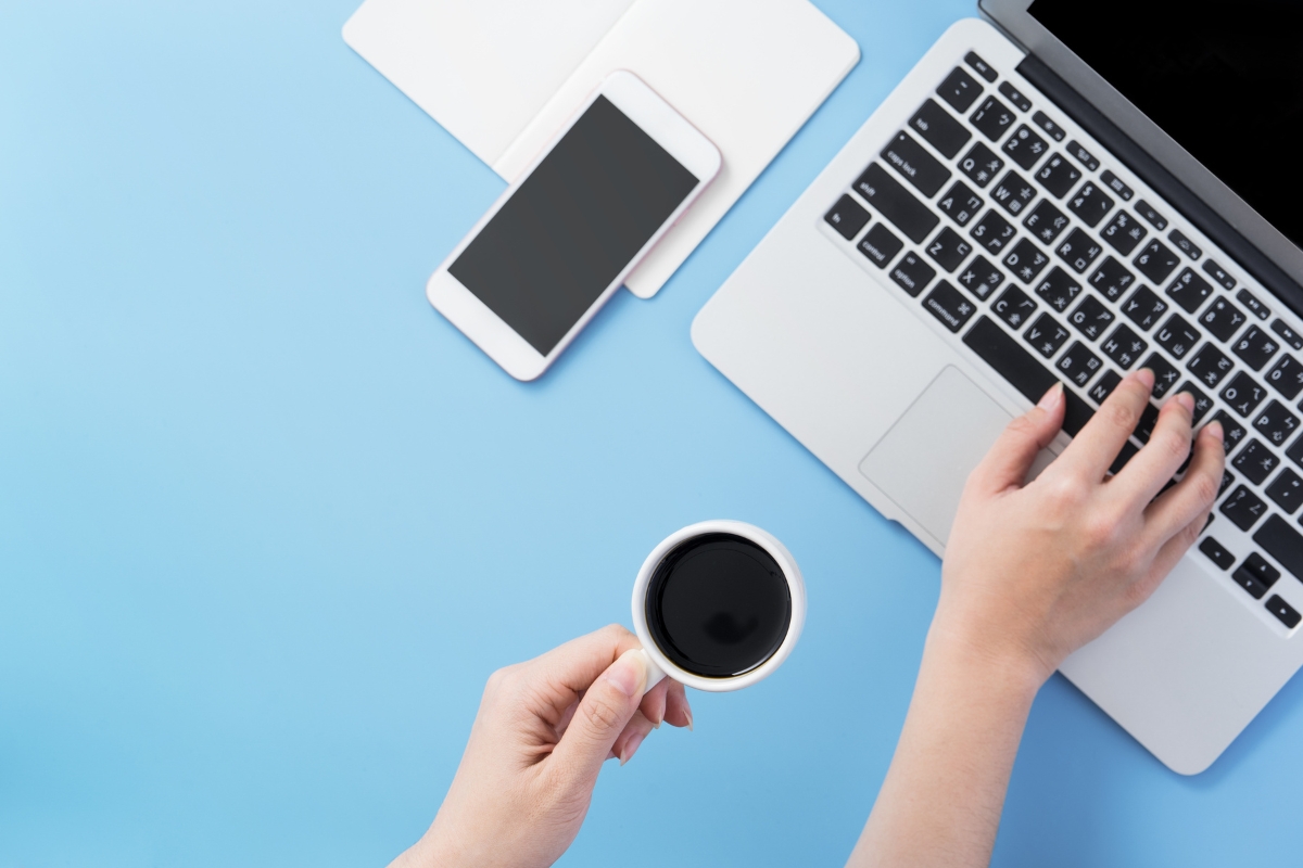 A person holds a cup of black coffee while typing on a laptop. A smartphone and a tablet are placed on the light blue desk, with an open browser tab asking, "What is UX writing?