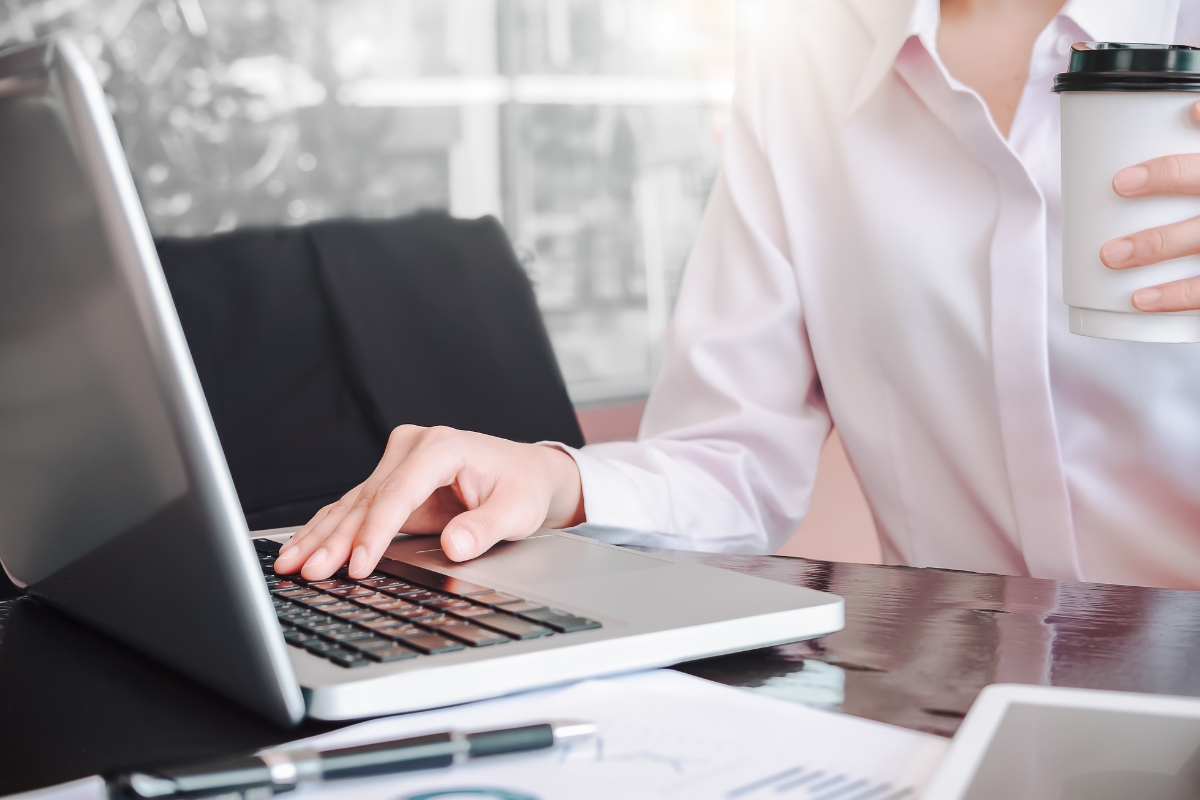 A person in a white shirt is working on a laptop while holding a coffee cup, possibly researching what is UX writing. Papers and a pen are on the desk.