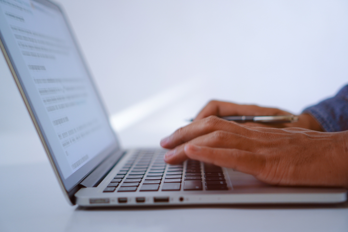 Close-up of hands typing on a laptop keyboard with a document open on the screen, titled "What is UX Writing?" The person holds a pen in their left hand.