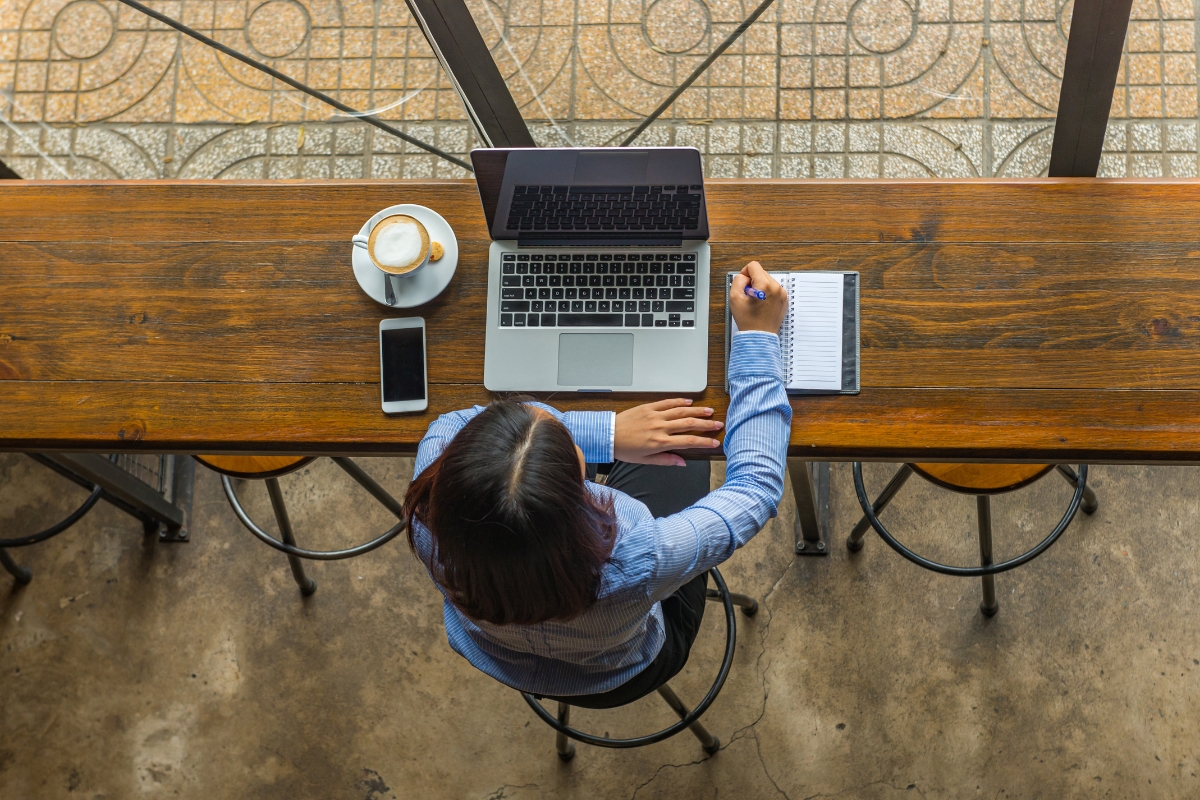 A person sits at a wooden counter working on a laptop with a phone, notepad, and cup of coffee on the table. The scene is viewed from above, perhaps pondering what is UX writing as they type away.