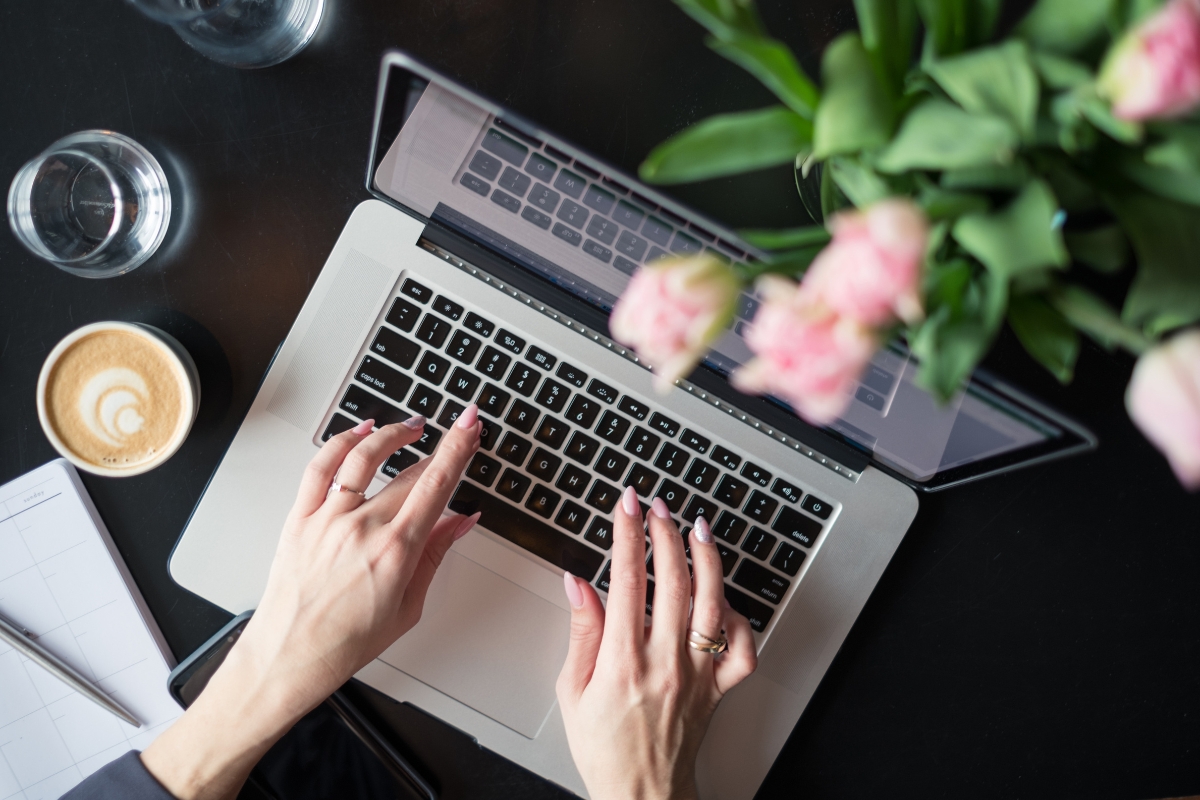 Top view of a person typing on a laptop keyboard, with a cup of coffee, a glass of water, a notepad, a pen labeled "what is UX writing," and a bouquet of pink flowers on the table.