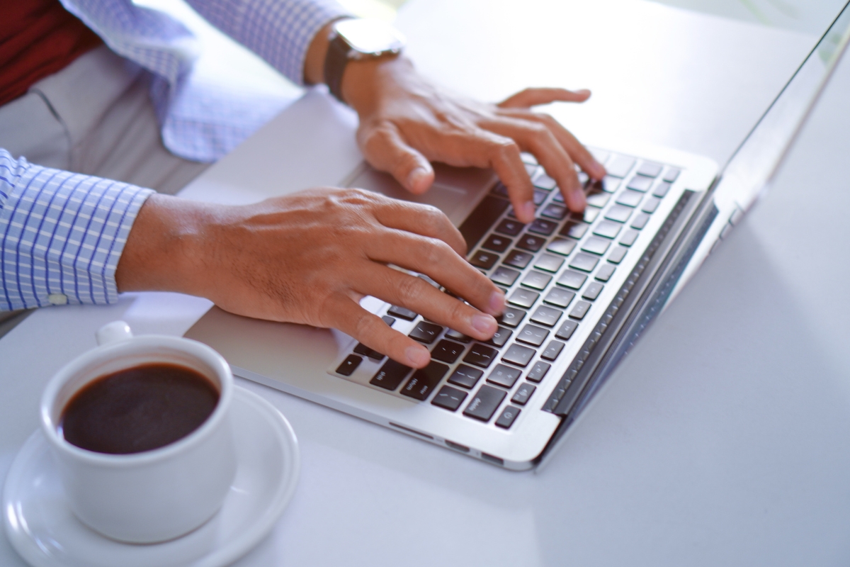 A person types on a laptop keyboard while seated at a desk with a cup of coffee nearby, perhaps wondering, "What is UX writing?