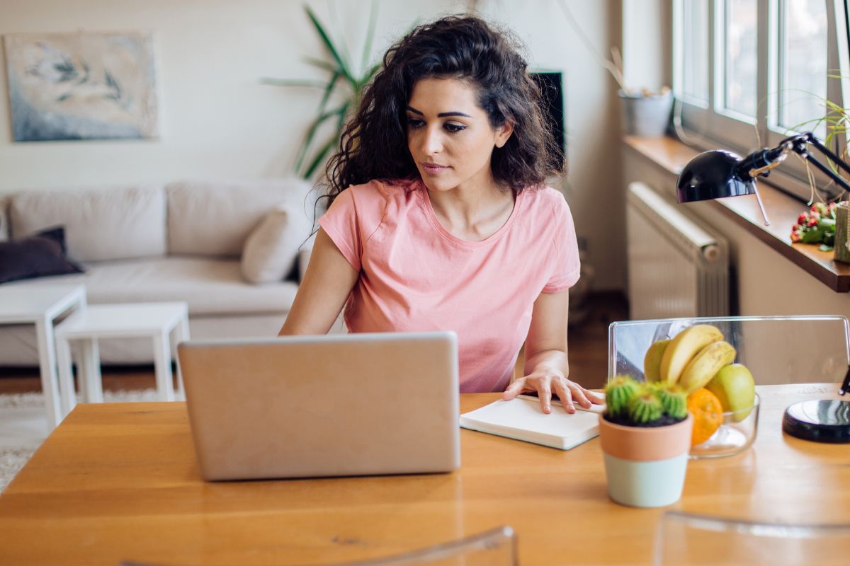 Person with long curly hair in a pink shirt working on a laptop at a wooden table with a notepad and a bowl of fruits nearby, possibly researching What Are SERPs. There is a couch and indoor plants in the background.