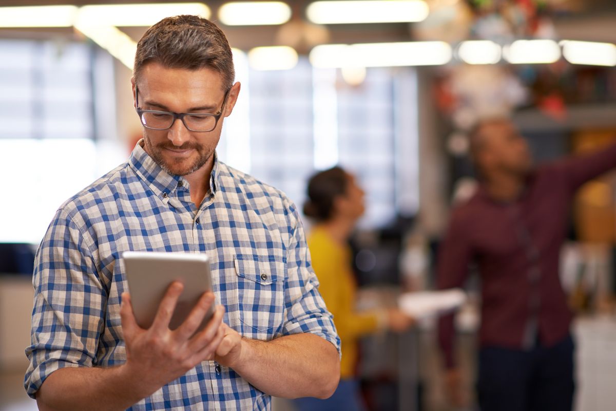 A man in a plaid shirt and glasses looks at a tablet in an office setting with two blurred figures working in the background, possibly researching What Are SERPs.