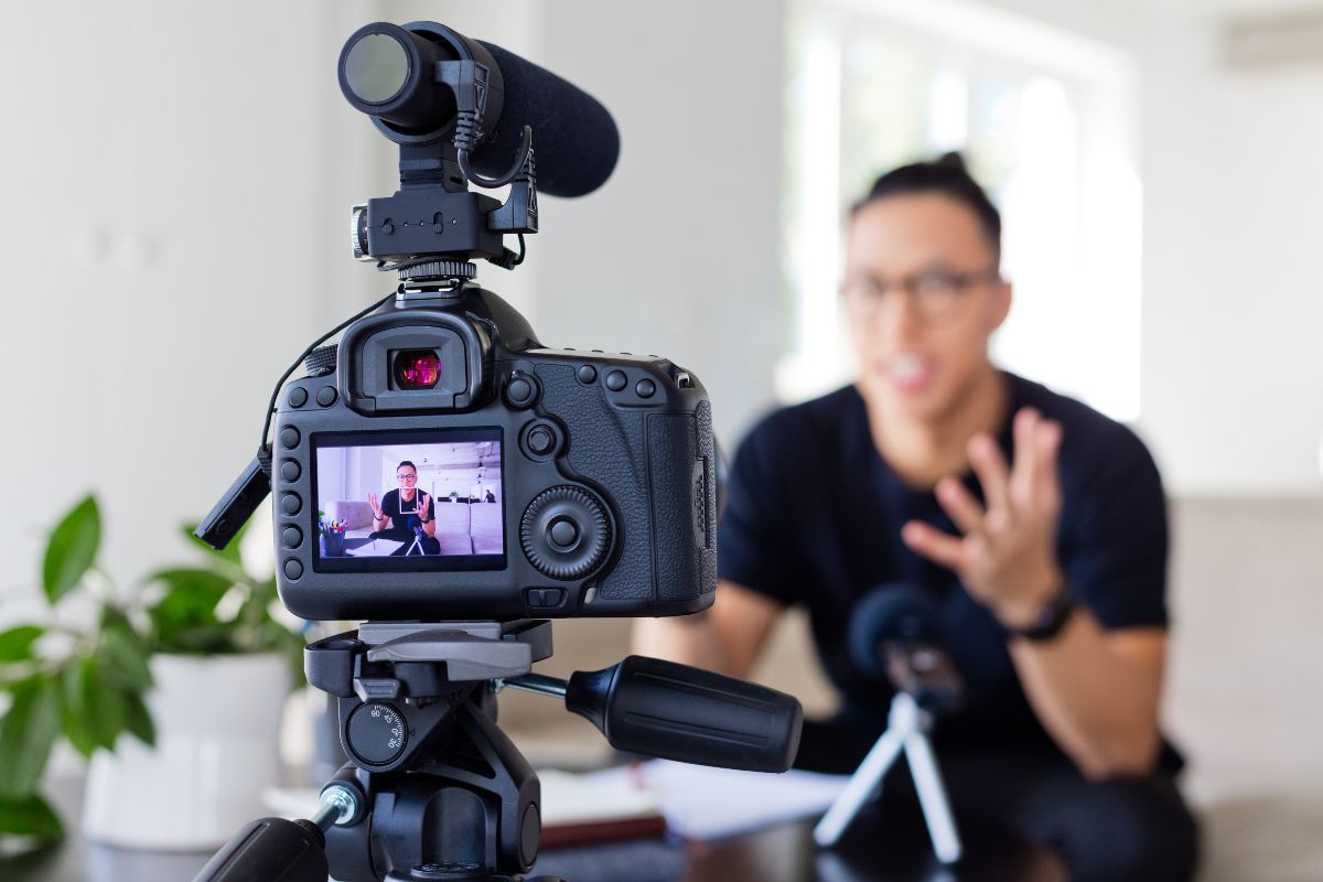 A person is giving video testimonials in front of a camera mounted on a tripod, with a microphone attached and a plant in the background.